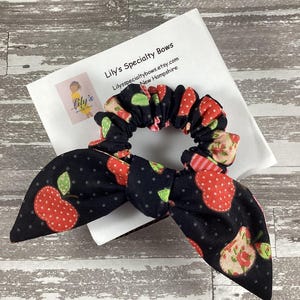 May include: A black scrunchie with a red and white apple print. The scrunchie is tied in a bow and has a white background with green leaves. The scrunchie is on a white card with the text "Lily's Specialty Bows" and "Lilyspecialtybows.etsy.com New Hampshire".
