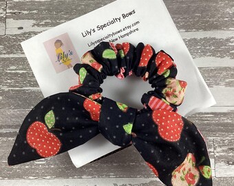 May include: A black scrunchie with a red and white apple print. The scrunchie is tied in a bow and has a white background with green leaves. The scrunchie is on a white card with the text "Lily's Specialty Bows" and "Lilyspecialtybows.etsy.com New Hampshire".