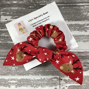 May include: Red and white scrunchie with a reindeer and star pattern. The scrunchie is tied in a bow and is on a white background with text that reads "Lily's Specialty Bows Lilysspecialtybows.etsy.com Made in New Hampshire".
