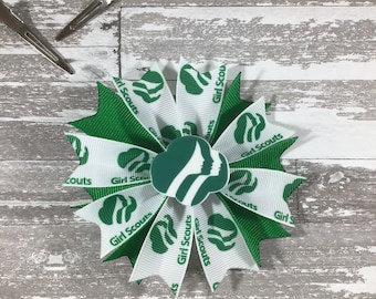May include: A green and white bow with the words "Girl Scouts" printed on it. The bow has a green Girl Scout logo in the center.