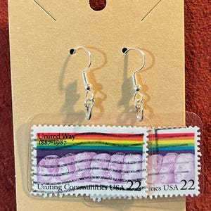 May include: Silver-tone dangle earrings featuring United Way postage stamp designs. The stamps display a rainbow over a group of faces, with text reading "United Way 1887-1987" and "Uniting Communities USA 22". The earrings are attached to a brown cardboard display.