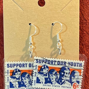 May include: Silver-tone dangle earrings featuring a vintage postage stamp design. The stamps display the text "SUPPORT OUR YOUTH" and images of children and adults. The earrings are attached to a brown card.