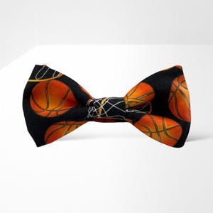 Puede incluir: Una pajarita negra con un patrón de balones de baloncesto naranjas. La pajarita está hecha de tela y tiene una forma clásica de pajarita.