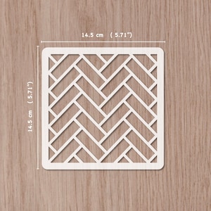 Puede incluir: Un esténcil cuadrado blanco con un diseño de espiga, que mide 14,5 cm (5,71 pulgadas) por lado. El diseño está recortado, creando un espacio negativo. El esténcil está colocado sobre una superficie de madera marrón claro.