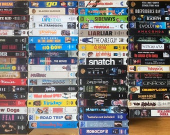 May include: A large collection of rectangular VHS movie tapes arranged in four vertical stacks. Each tape features a unique spine with printed film titles such as Point Break, Die Hard 2, The Karate Kid, Snatch, Heat, and Robocop 2 in various colors.
