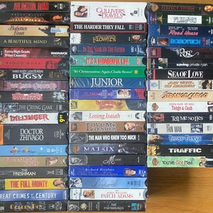 May include: A collection of VHS tapes, featuring titles like "Arlington Road," "Armageddon," and "Gulliver's Travels." The tapes are stacked, showcasing a variety of movie genres and cover art designs. The collection includes titles from the 1990s.