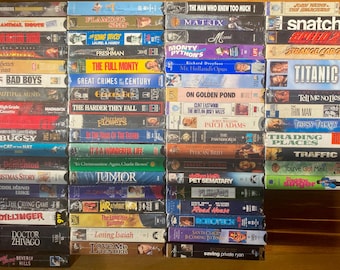 May include: A collection of VHS tapes, stacked on a wooden surface. Titles include "Titanic", "Bad Boys", "The Matrix", and "Snatch". The tapes are in various colors and designs, showcasing a variety of movie genres.