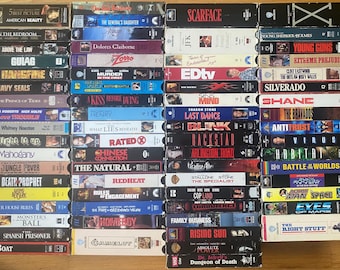 May include: A collection of numerous VHS tapes arranged in four vertical columns. Each tape features a unique spine with distinct movie titles, including Scarface, JFK, and American Beauty, displayed in various fonts, colors, and graphic designs.