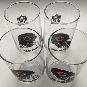 May include: Four clear glass tumblers with the Chicago Bears football team logo. The logo features a navy blue helmet with an orange C and the word "BEARS" in white. The NFL logo is also visible on the glasses.