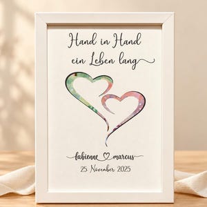 Könnte beinhalten: Ein weißer Bilderrahmen mit dem deutschen Text "Hand in Hand ein Leben lang" über zwei ineinander verschlungenen Herzausschnitten, die einen bunten Hintergrund zeigen. Darunter stehen die Namen "fabienne" und "marcus" sowie das Datum "25. November 2025" in schwarzer Schrift.