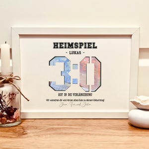Könnte beinhalten: Ein weißer gerahmter Druck mit dem Text "HEIMSPIEL - LUKAS - 3:0" in Schwarz. Die Zahlen sind mit einer Collage aus farbenfrohen Geldscheinen gefüllt. Darunter steht der Text "AUF IN DIE VERLÄNGERUNG!" und eine Geburtstagsbotschaft.
