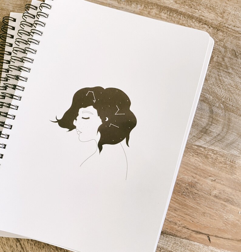 May include: A white spiral-bound notebook with a black and white illustration of a woman's profile. The woman's hair is filled with constellations and a crescent moon. The notebook is open on a wooden surface.