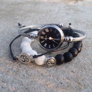 May include: A set of three bracelets featuring a silver watch with Roman numeral markings, a white beaded bracelet with an Om symbol charm, and a black beaded bracelet with a yin yang symbol charm.