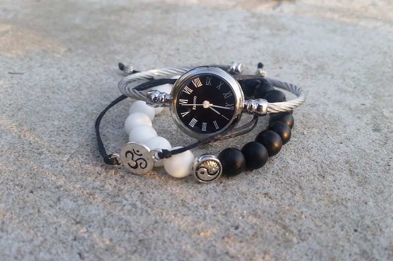 May include: A set of three bracelets featuring a silver watch with Roman numeral markings, a white beaded bracelet with an Om symbol charm, and a black beaded bracelet with a yin yang symbol charm.