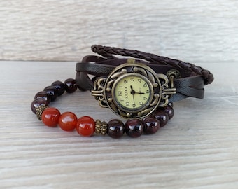 May include: A vintage-style watch with a cream-colored face and bronze-toned casing. The watch is paired with a dark brown leather wrap-around strap and a beaded bracelet with red and dark brown beads. The watch face reads "O.L.SEA QUARTZ".