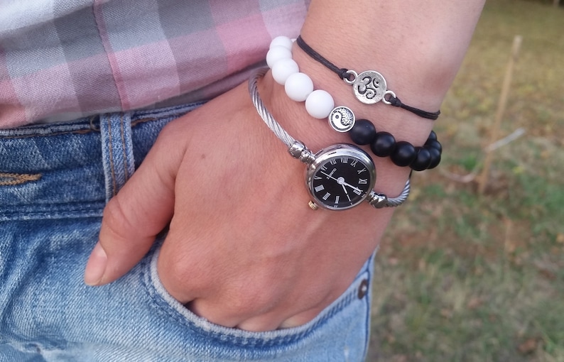 May include: A silver bangle bracelet with a black face watch with Roman numeral markings. The watch is worn with a black beaded bracelet and a white beaded bracelet with a silver Om charm.