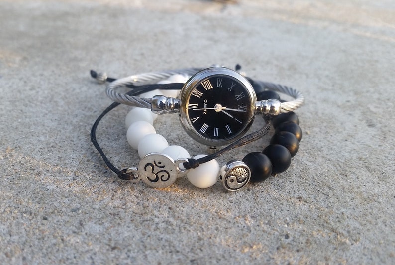 May include: A set of three bracelets featuring black and white beads, a silver bangle, and a silver watch with a black face and Roman numeral markings. The watch face reads "Quartz".