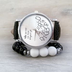 May include: A silver wristwatch with a black leather band and white face. The face has black numbers and the text "who cares i'm late anyways". The watch is on a white surface with black and white beads.