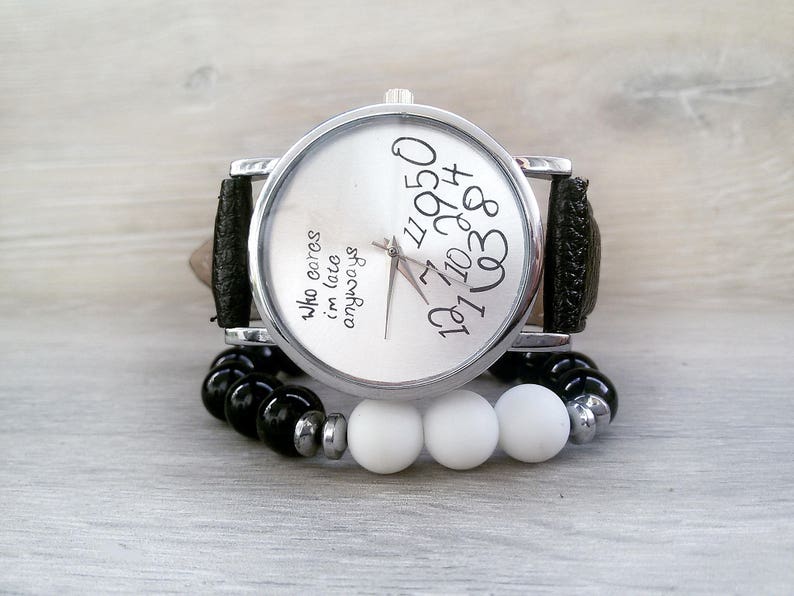 May include: A silver wristwatch with a black leather band and white face. The face has black numbers and the text "who cares i'm late anyways". The watch is on a white surface with black and white beads.