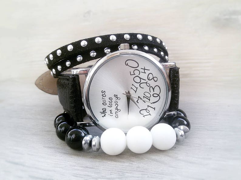 May include: A silver wristwatch with a black leather band and a white face. The face has black numbers and the text "Who cares I'm late anyways". The watch is wrapped in a black studded band.
