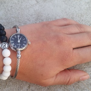 May include: A silver bangle watch with a black face and Roman numeral hour markers. The watch is worn on a wrist with two bracelets, one with black beads and one with white beads. The bracelets also feature a silver charm with an Om symbol and a yin yang symbol.