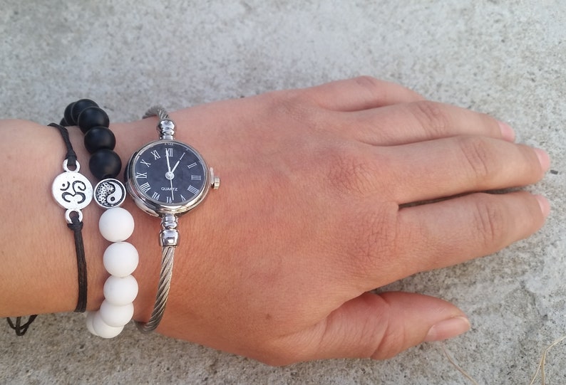 May include: A silver bangle watch with a black face and Roman numeral hour markers. The watch is worn on a wrist with two bracelets, one with black beads and one with white beads. The bracelets also feature a silver charm with an Om symbol and a yin yang symbol.