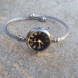 May include: A silver bangle watch with a black face and Roman numeral hour markers. The watch has a quartz movement.