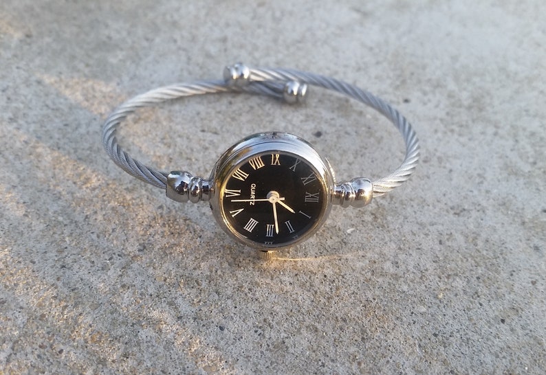May include: A silver bangle watch with a black face and Roman numeral hour markers. The watch has a quartz movement.