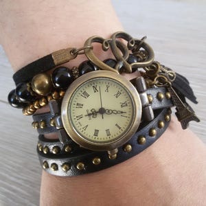 May include: A vintage-style watch with multiple layers. The watch has a cream-coloured face with Roman numerals and a bronze-coloured case. It's attached to a black leather strap with gold-coloured studs and a black suede band.