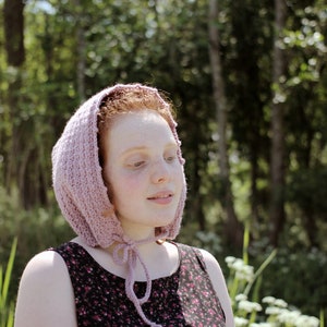 May include: A young person with red hair wearing a pink knitted hooded hat and a floral print dress. The person is standing in a forest setting.