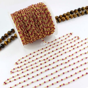 Puede incluir: Un carrete de cadena chapada en oro con pequeñas cuentas facetadas de color rojo rubí. La cadena está dispuesta en filas sobre una superficie blanca.