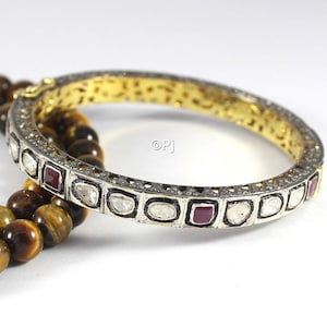 May include: A gold bangle bracelet with a silver band set with small diamonds and red gemstones. The bracelet is on top of a brown tiger's eye beaded necklace.