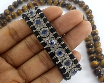 May include: A black cord bracelet with a silver chain and blue and white gemstones. The bracelet is on a hand with a tiger's eye beaded necklace in the background.