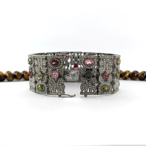 May include: A silver bracelet with a floral design and a variety of colorful gemstones, including pink, green, and red. The bracelet is open and the clasp is visible.