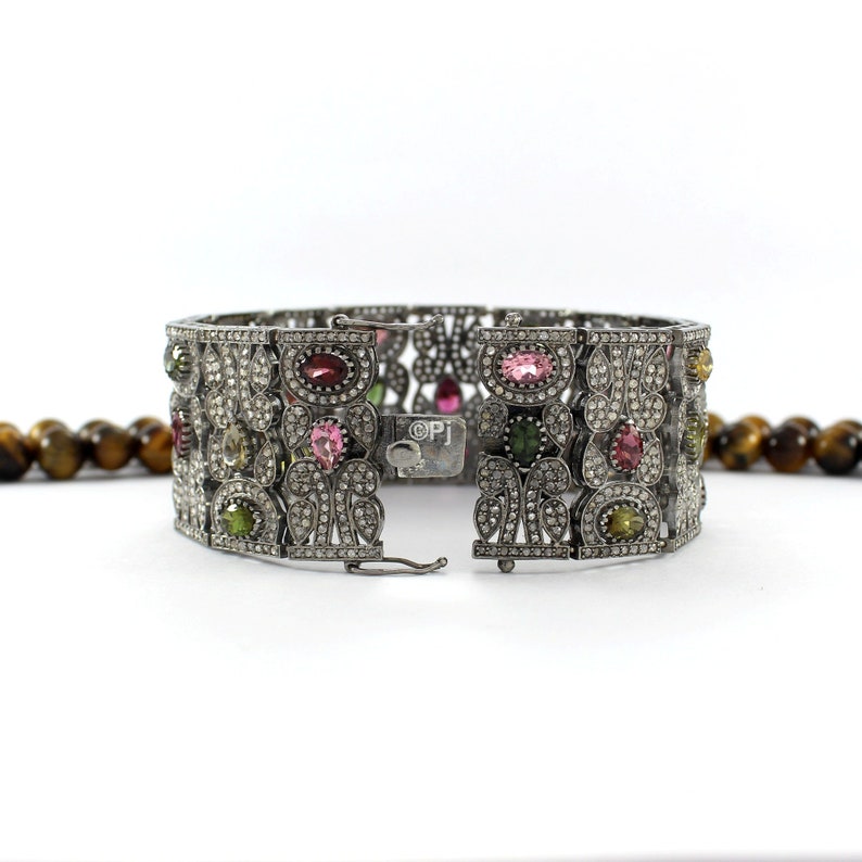 May include: A silver bracelet with a floral design and a variety of colorful gemstones, including pink, green, and red. The bracelet is open and the clasp is visible.