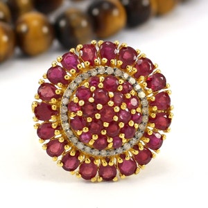 Op de afbeelding: Een gouden ring met een grote, ronde middelste steen die omringd is door kleinere stenen. De middelste steen is een robijn en de kleinere stenen zijn diamanten. De ring is gezet in een gouden band.