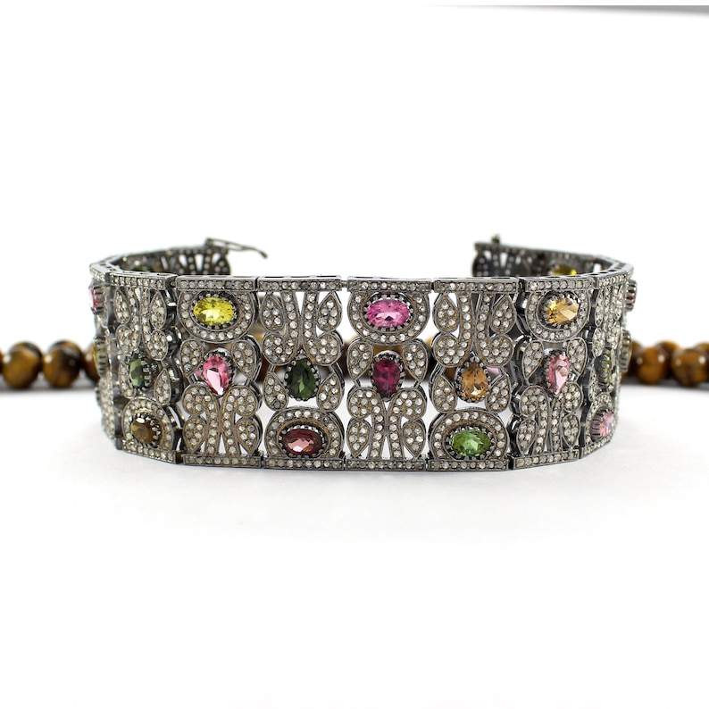 May include: A silver bracelet with a geometric design featuring a variety of colorful gemstones, including pink, green, and yellow.