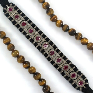 May include: A black velvet bracelet with a silver and diamond design. The bracelet is adorned with red ruby stones. The bracelet is paired with a brown tiger's eye bead necklace.