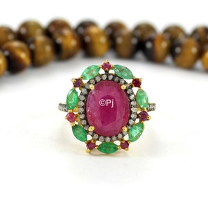 Könnte beinhalten: Ein goldener Ring mit einem großen ovalen Rubin, umgeben von kleineren Smaragden und Diamanten. Der Ring ist in einem floralen Design gefasst.