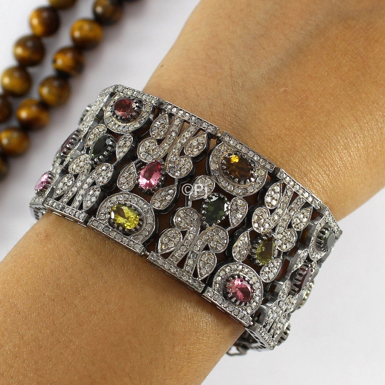 May include: A silver bracelet with a floral design and colorful gemstones. The bracelet is set with small white diamonds and features pink, green, and yellow gemstones.