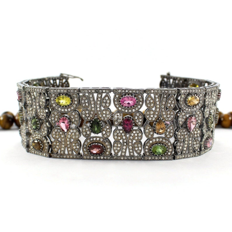 May include: A silver bracelet with a delicate filigree design and colorful gemstones. The bracelet features a variety of oval-shaped gemstones in shades of pink, green, and yellow, set in a silver frame. The bracelet is adorned with small, sparkling diamonds.