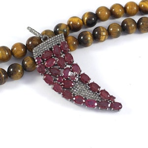 May include: A silver pendant with a horn shape, encrusted with red ruby and clear diamond stones. The pendant is displayed on a brown tiger's eye beaded necklace.