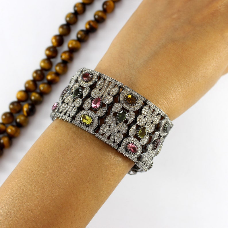 May include: A wide silver bracelet with a floral design and colorful gemstones. The bracelet is set with small diamonds and features a variety of colors, including pink, green, and yellow.