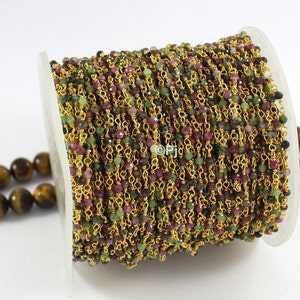 Puede incluir: Un carrete de cadena dorada con cuentas de piedras preciosas verdes, rosas y negras. La cadena está hecha de pequeños anillos metálicos unidos.