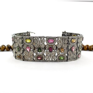 May include: A silver bracelet with a detailed design featuring a variety of colorful gemstones, including pink, green, and yellow.