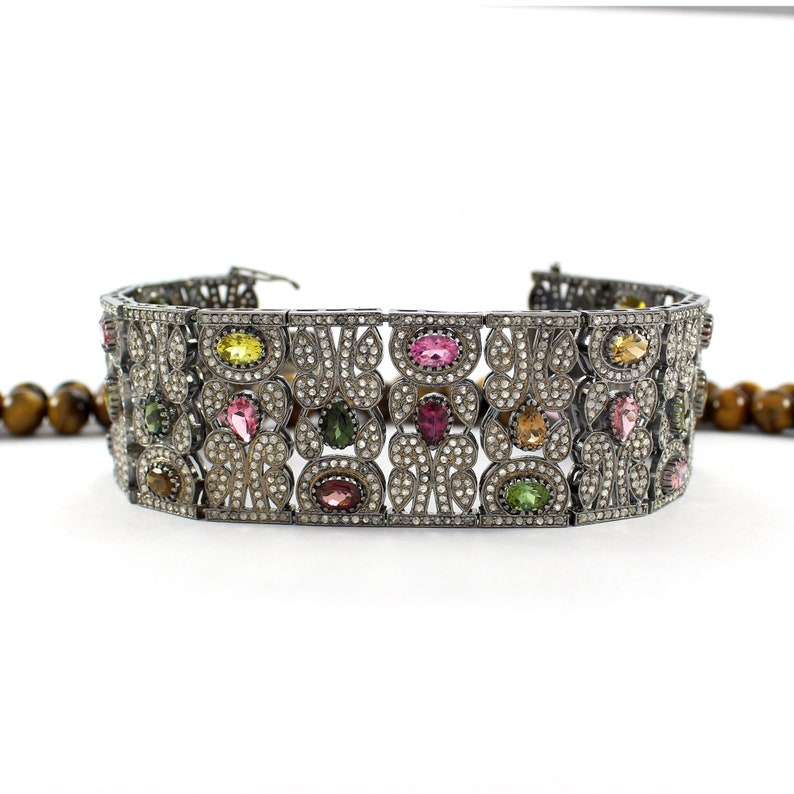 May include: A silver bracelet with a detailed design featuring a variety of colorful gemstones, including pink, green, and yellow.