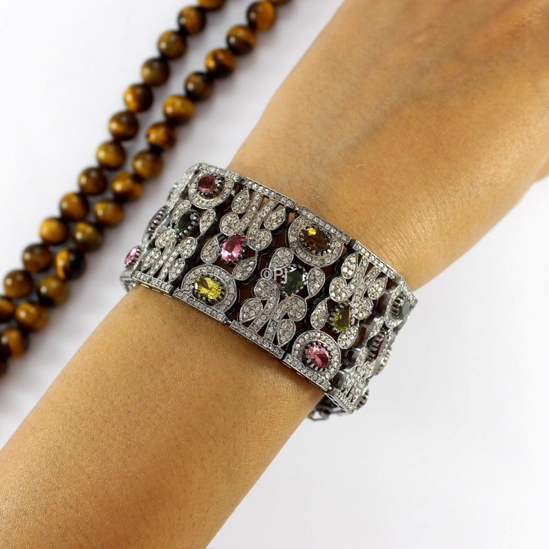 May include: A silver bangle bracelet with a floral design and colorful gemstones. The bracelet is set with small white diamonds and features pink, green, and yellow gemstones.