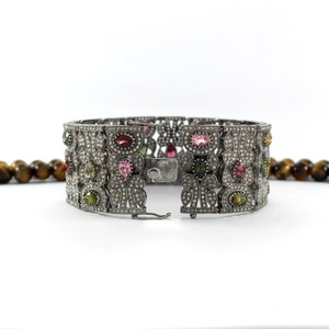 May include: A silver bracelet with a floral design and colorful gemstones. The bracelet is open and has a clasp.