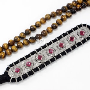 May include: A black velvet bracelet with a silver and diamond design. The bracelet is adorned with red gemstones. The bracelet is paired with a double strand of tiger's eye beads.