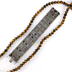 May include: A silver bracelet with a detailed design featuring colorful gemstones set in a silver filigree. The bracelet is paired with a brown tiger's eye bead necklace.